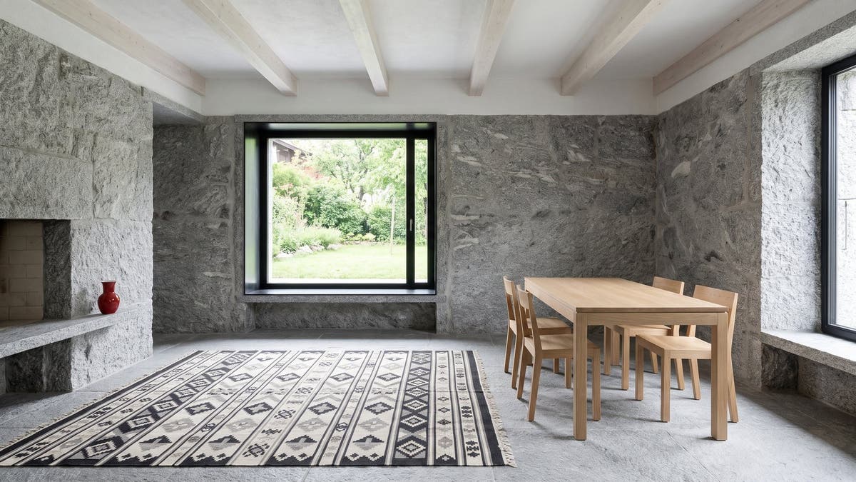 A restored Ticino stone house with granite walls, pale wooden beams and a black and ivory flatweave rug on a grey stone floor