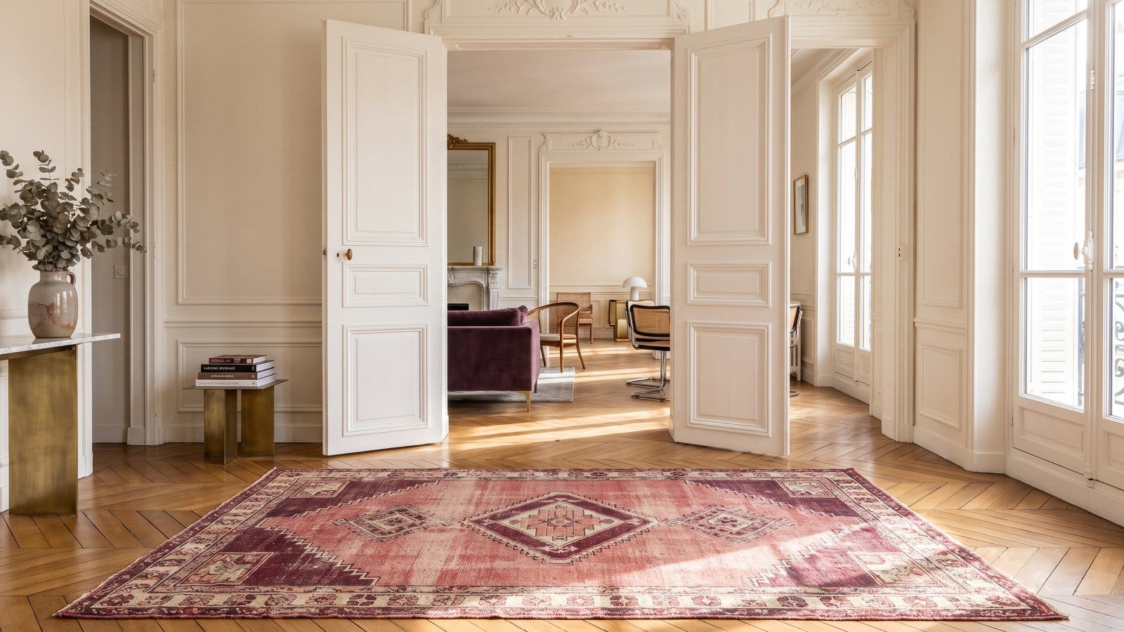 Parquet point de Hongrie baigné de lumière dans un appartement parisien, tapis vintage aux tons rose et bordeaux, et grandes portes d'époque ouvertes sur la pièce voisine