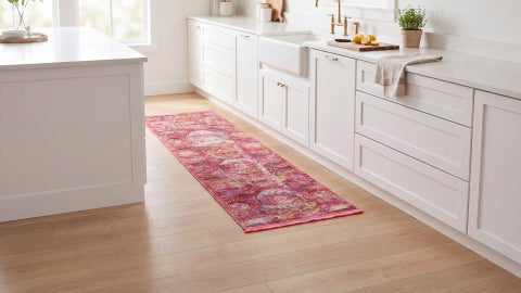Pink and red runner with a faded medallion pattern and fringe on a wood floor beside white kitchen cabinets