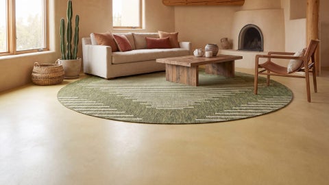 Round olive green rug with cream line diamond pattern under a wooden coffee table, in front of a beige sofa and a fireplace