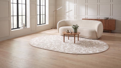 Oval rug with a beige and ivory floral vine pattern on a wood floor, under a small round table and cream curved sofa
