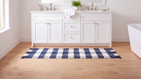 Navy and white buffalo check fringed runner on a wood floor in front of a white double sink vanity, with a tub beside it
