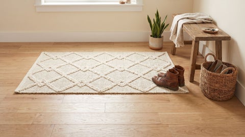 Cream textured rug with raised diamond lattice on a wood floor, brown leather boots on it, beside a bench and basket